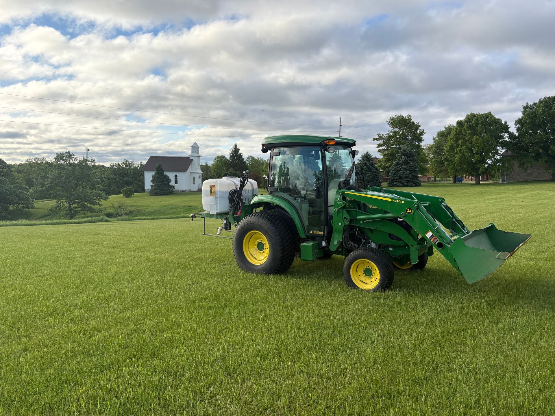 Green tractor mowing a grassy field under a cloudy sky, with a house and trees in the background.