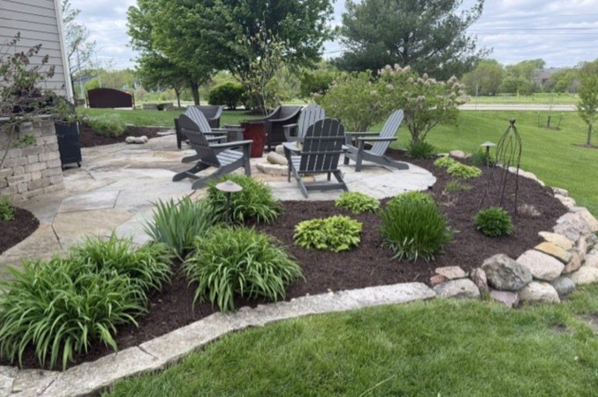 Backyard patio with black chairs, fire pit, and landscaped flower bed beside a grassy lawn.