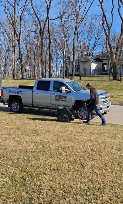 A person in a gray hoodie prepares a large piece of landscaping equipment parked behind a silver pickup truck on a lawn.