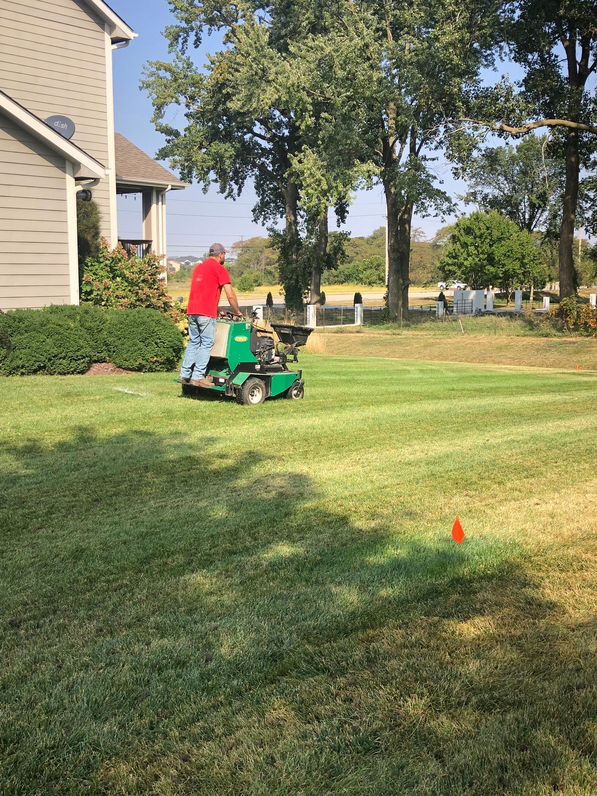 Person pushing a child on a green ride-on mower across a sunny suburban lawn.