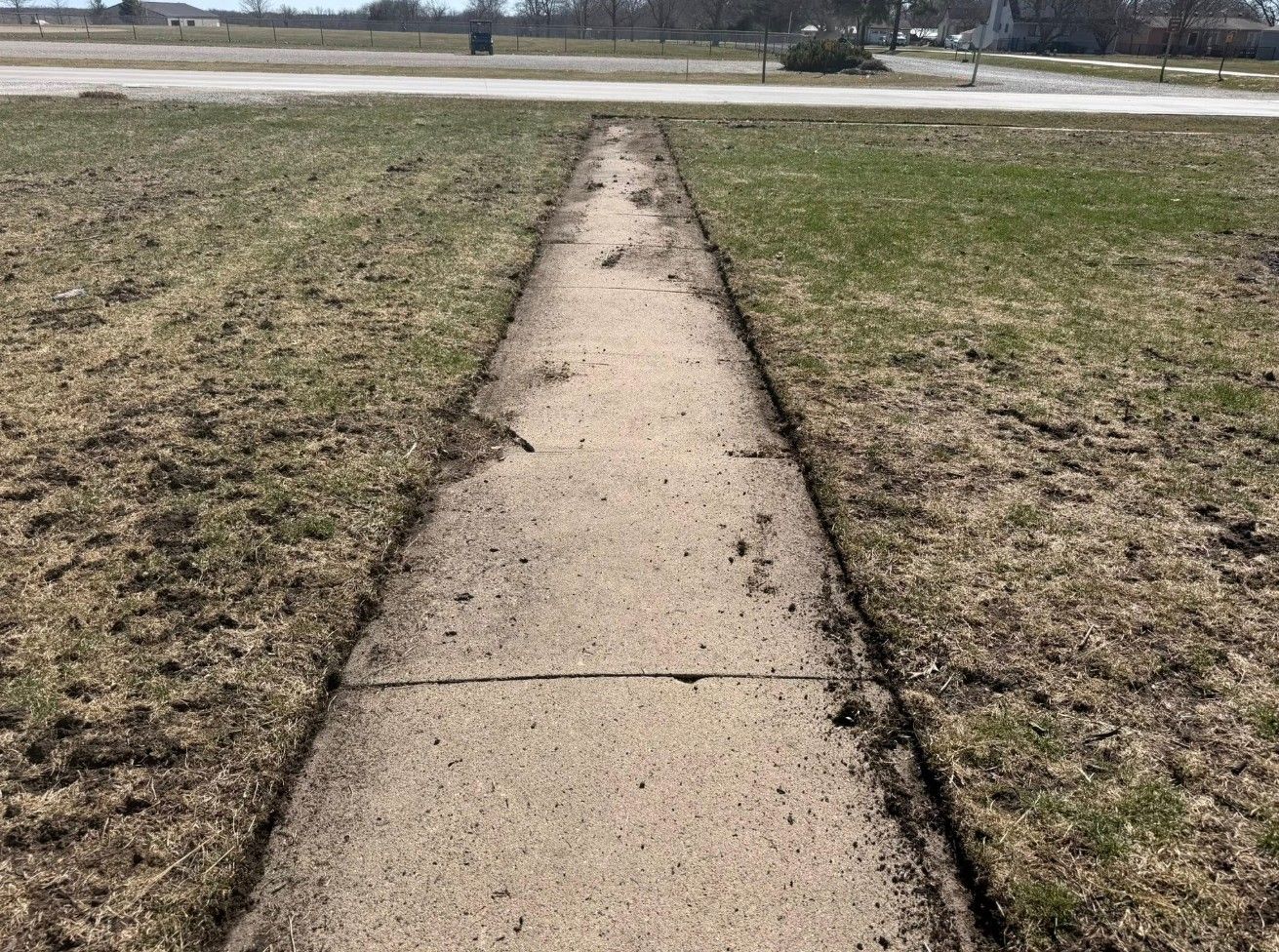 A straight, concrete sidewalk leads through a grassy field towards a road in the distance under a clear sky.