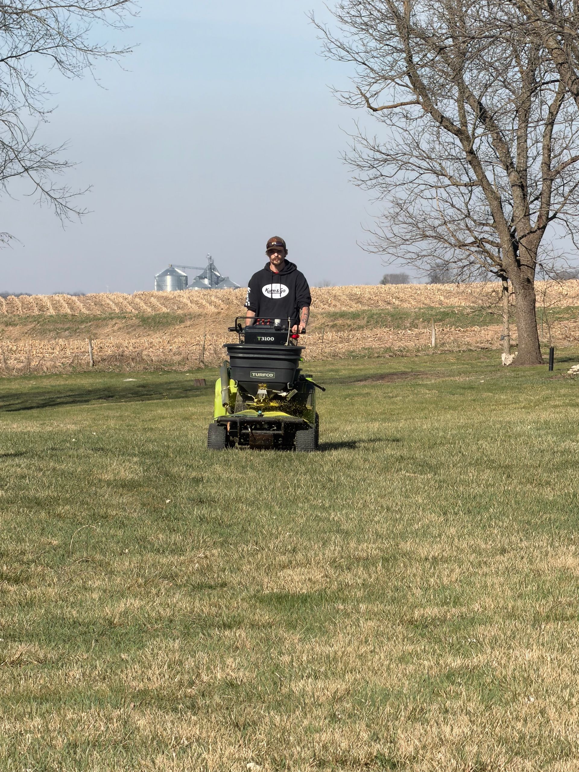 Person riding a green lawn mower across a grassy field with trees and a cornfield in the background