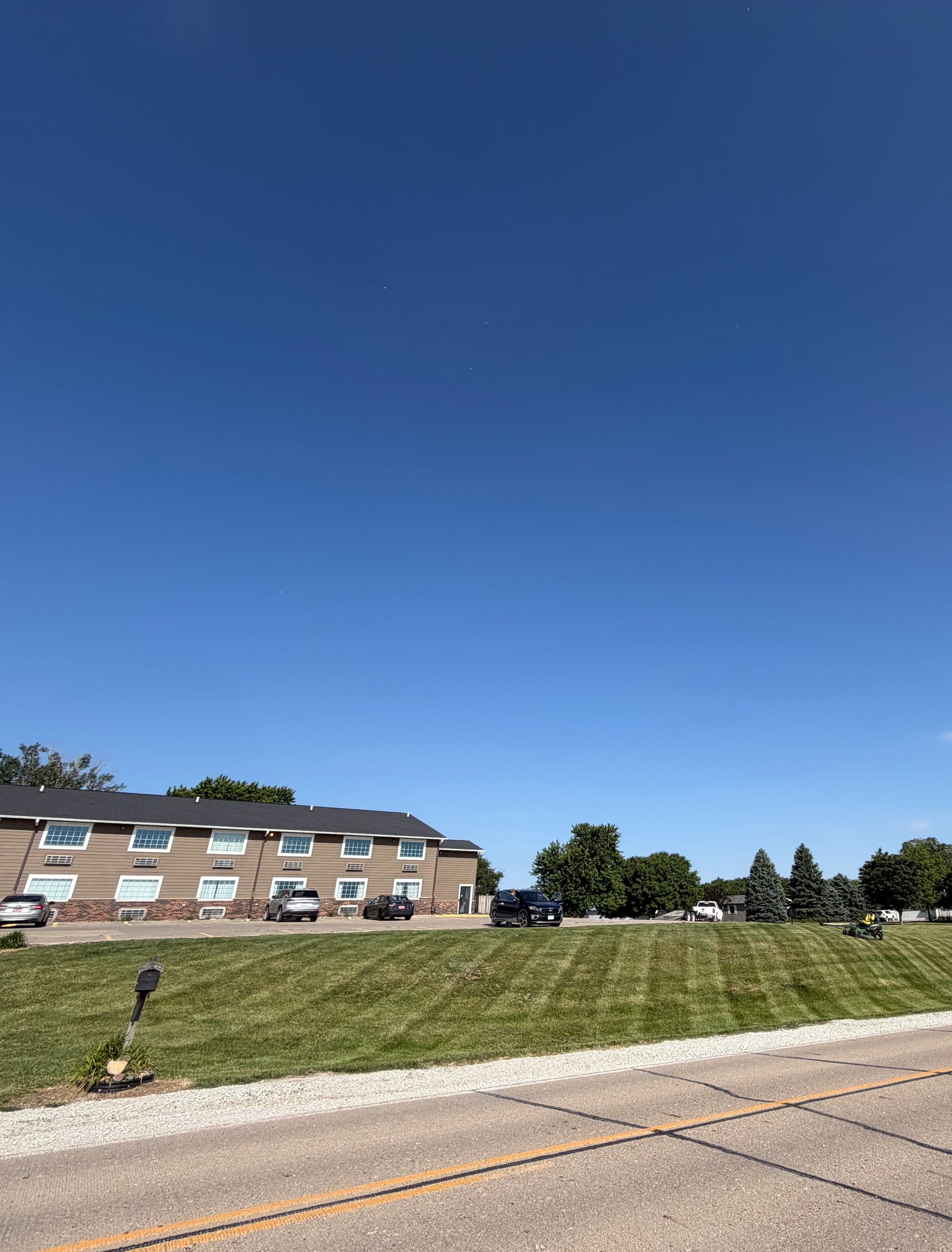 Brick apartment building beside a grassy slope under a clear blue sky, seen from a roadside