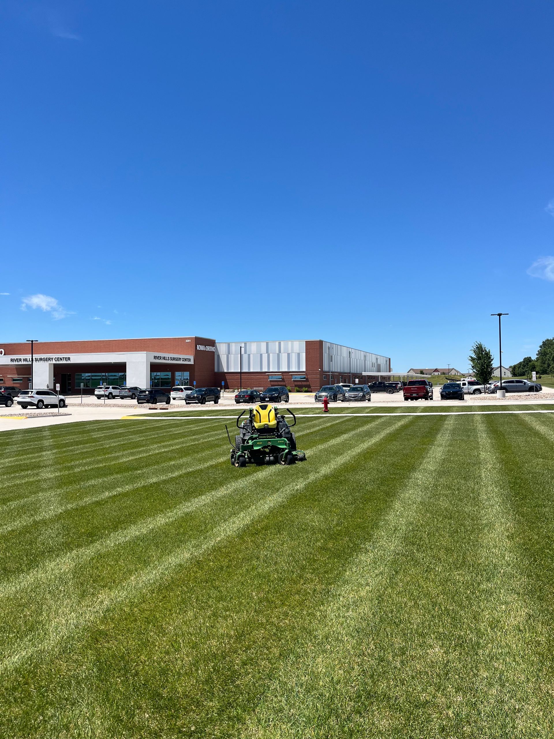 Person riding a green mower on a striped lawn in front of a brick building under a blue sky