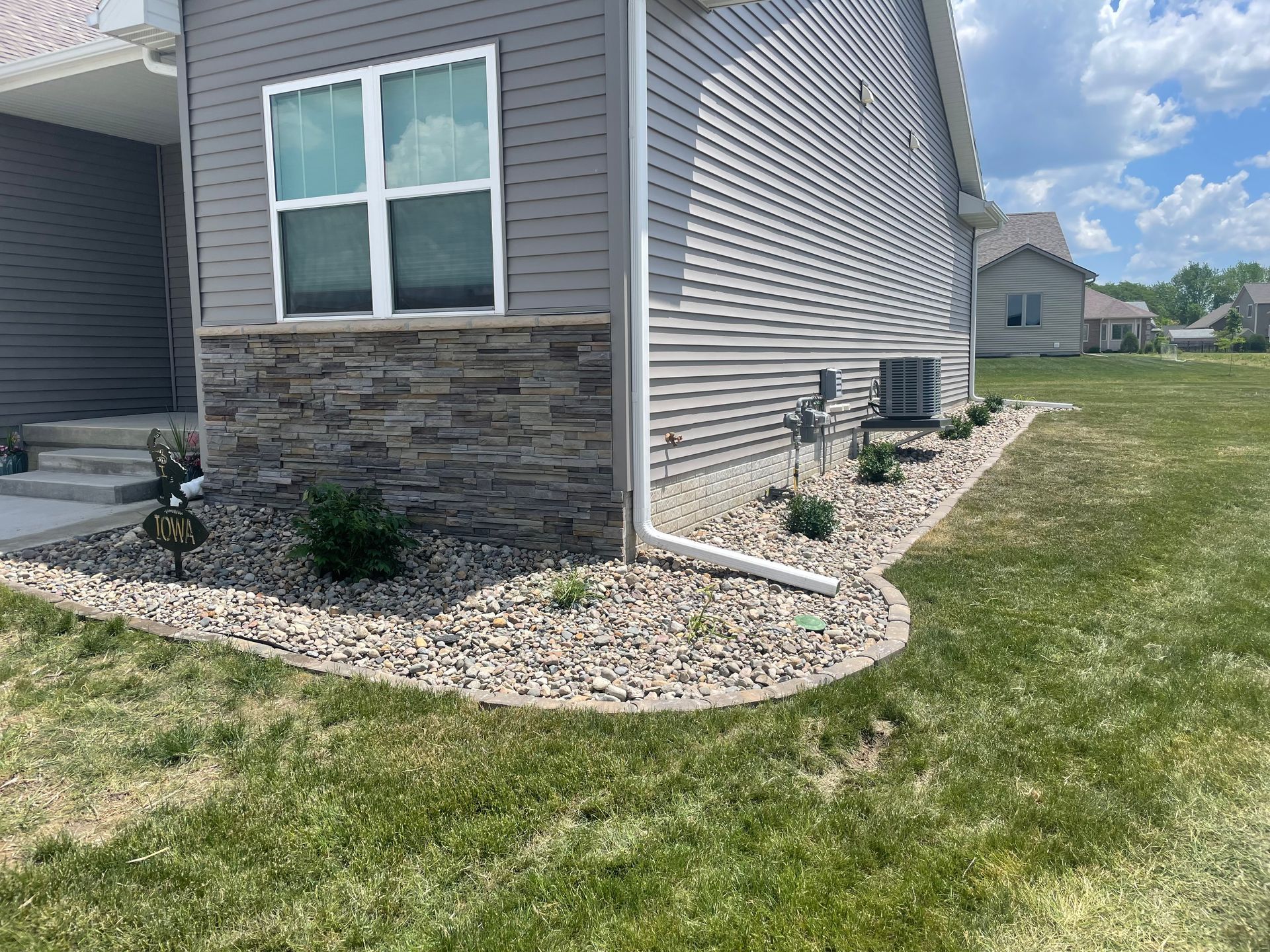 Corner of a gray house with stone siding, white-trimmed window, and landscaped rock border by a grassy yard