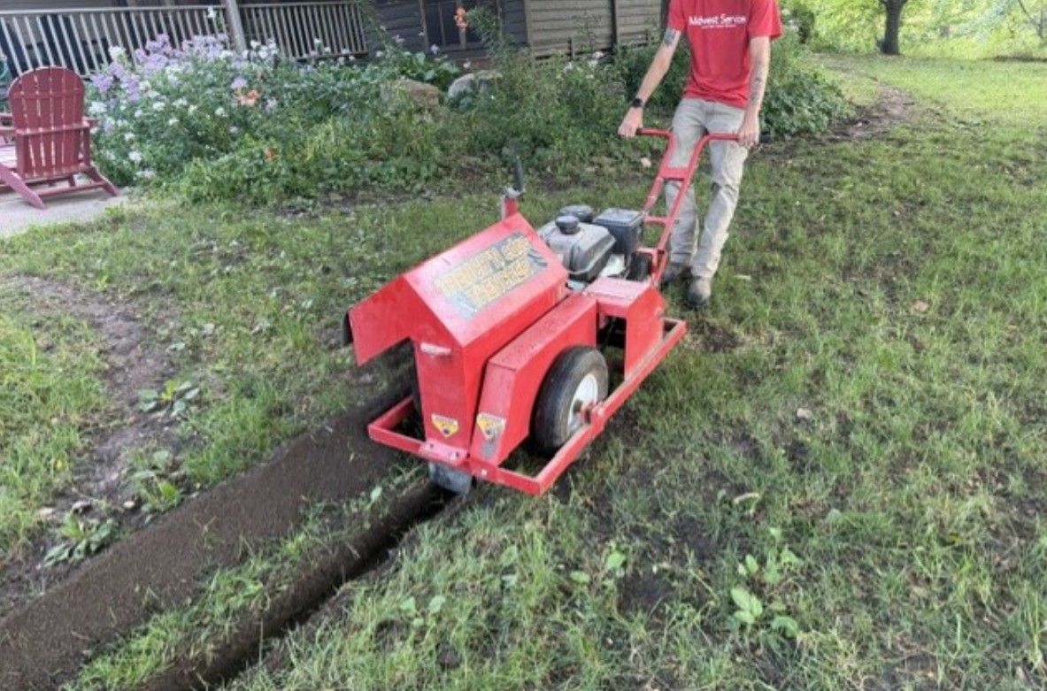 Worker operating a red trenching machine in a grassy yard, cutting a narrow trench.