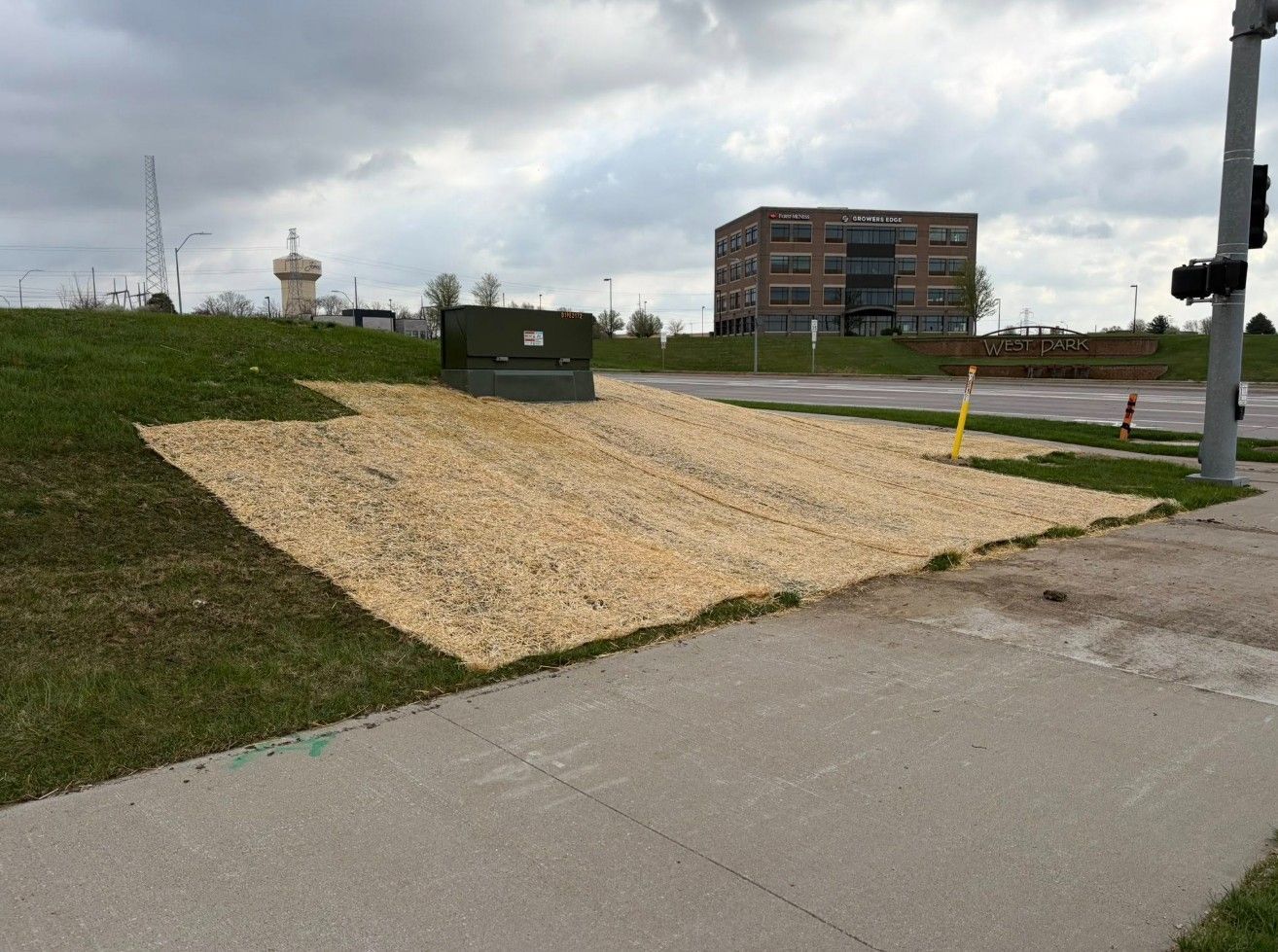 Gravel drainage ramp beside a paved path, with a utility box and apartment building under cloudy skies.