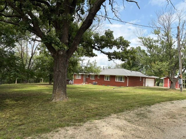 Red brick house with white trim under large trees beside a grassy yard and dirt driveway.