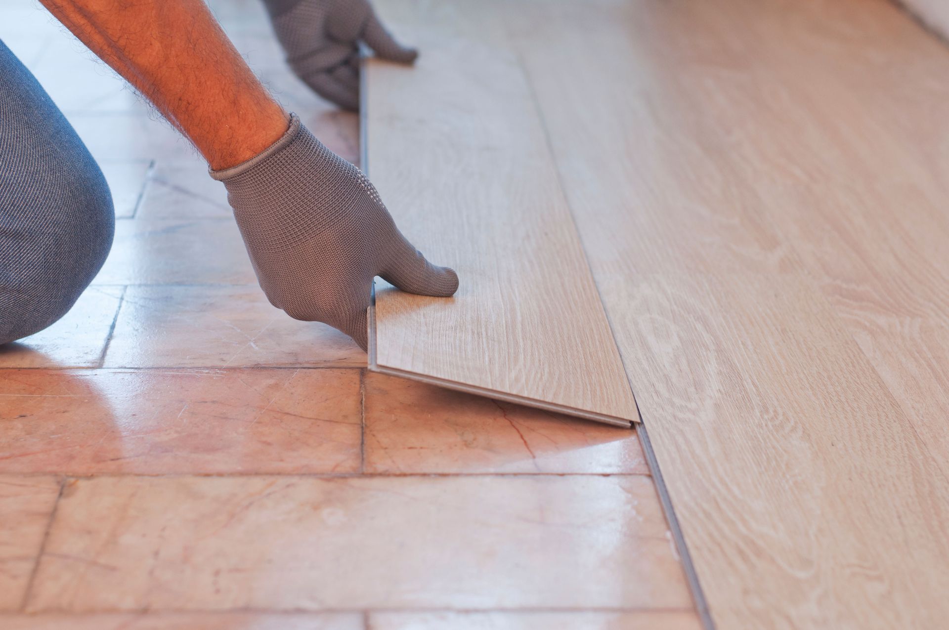 Person installing light-colored laminate flooring, wearing gloves, on a tiled surface.