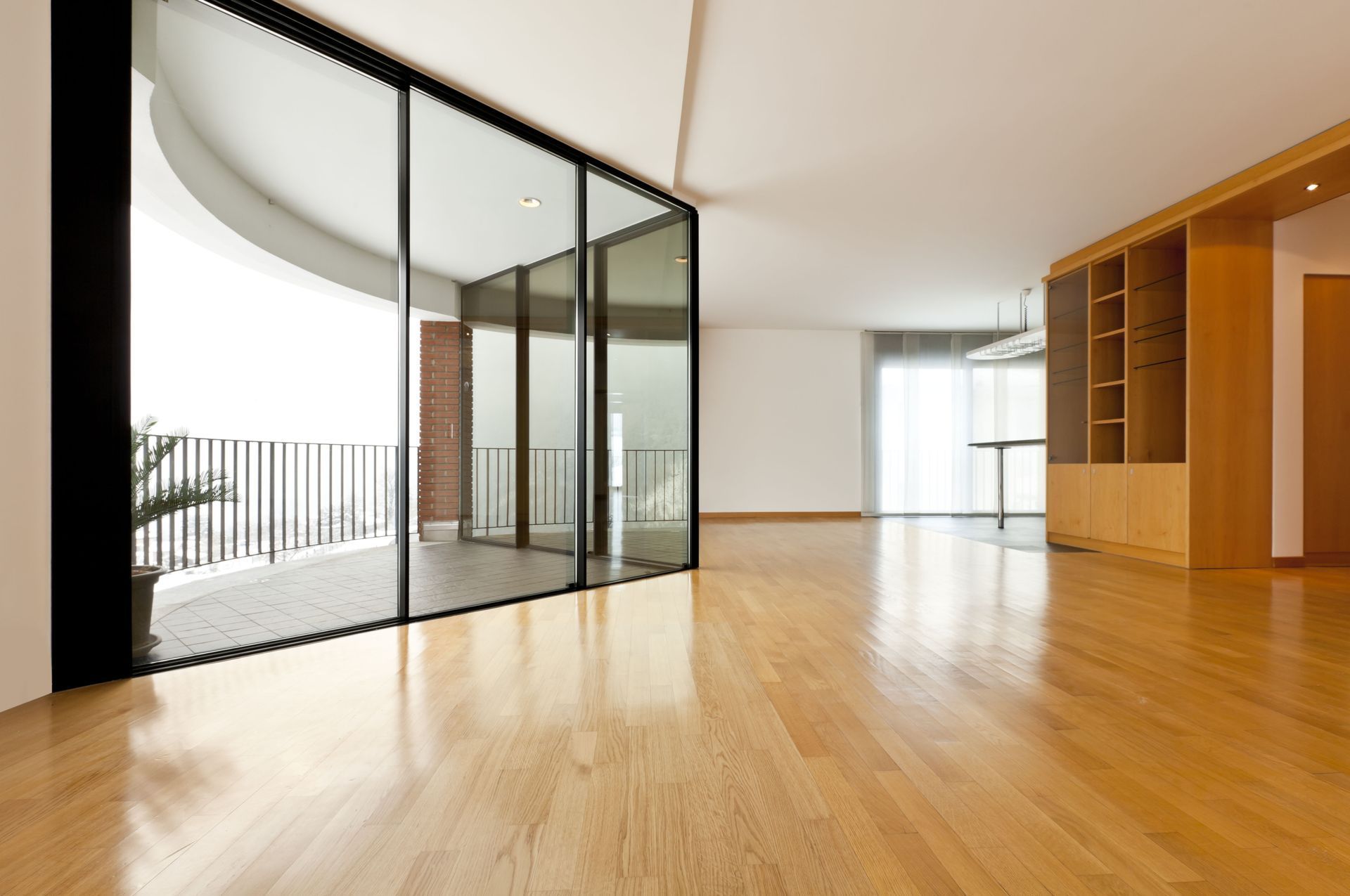 Empty modern room with wooden floor, large glass doors to a balcony, built-in shelving.