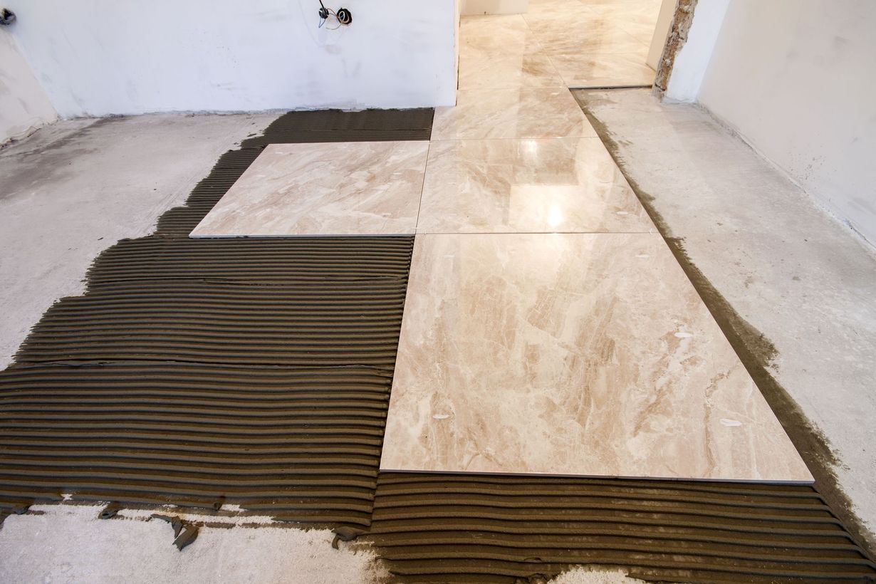 Floor tiles being installed, showing troweled mortar base with beige tiles in a room.