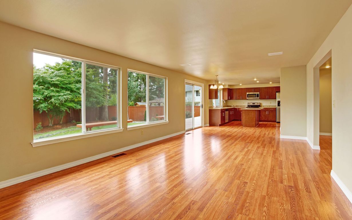 Empty living room with wood flooring, large windows, and an open kitchen with brown cabinets.