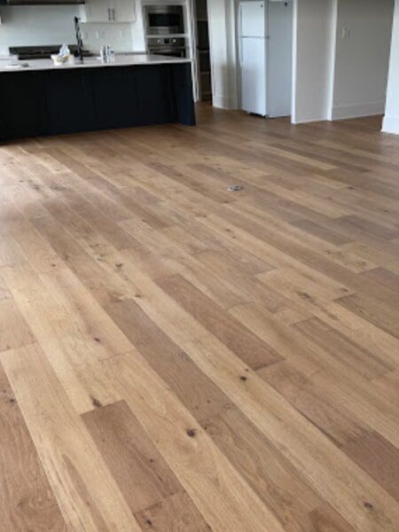 Light brown hardwood flooring in a kitchen, with a dark island and a white refrigerator in the background.