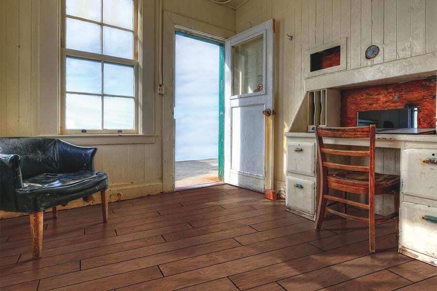 Interior of a weathered room with a desk, chair, and open doorway to a bright sky.