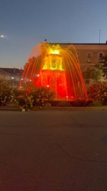 Lit fountain glowing red and yellow at dusk in a plaza, with a building in the background