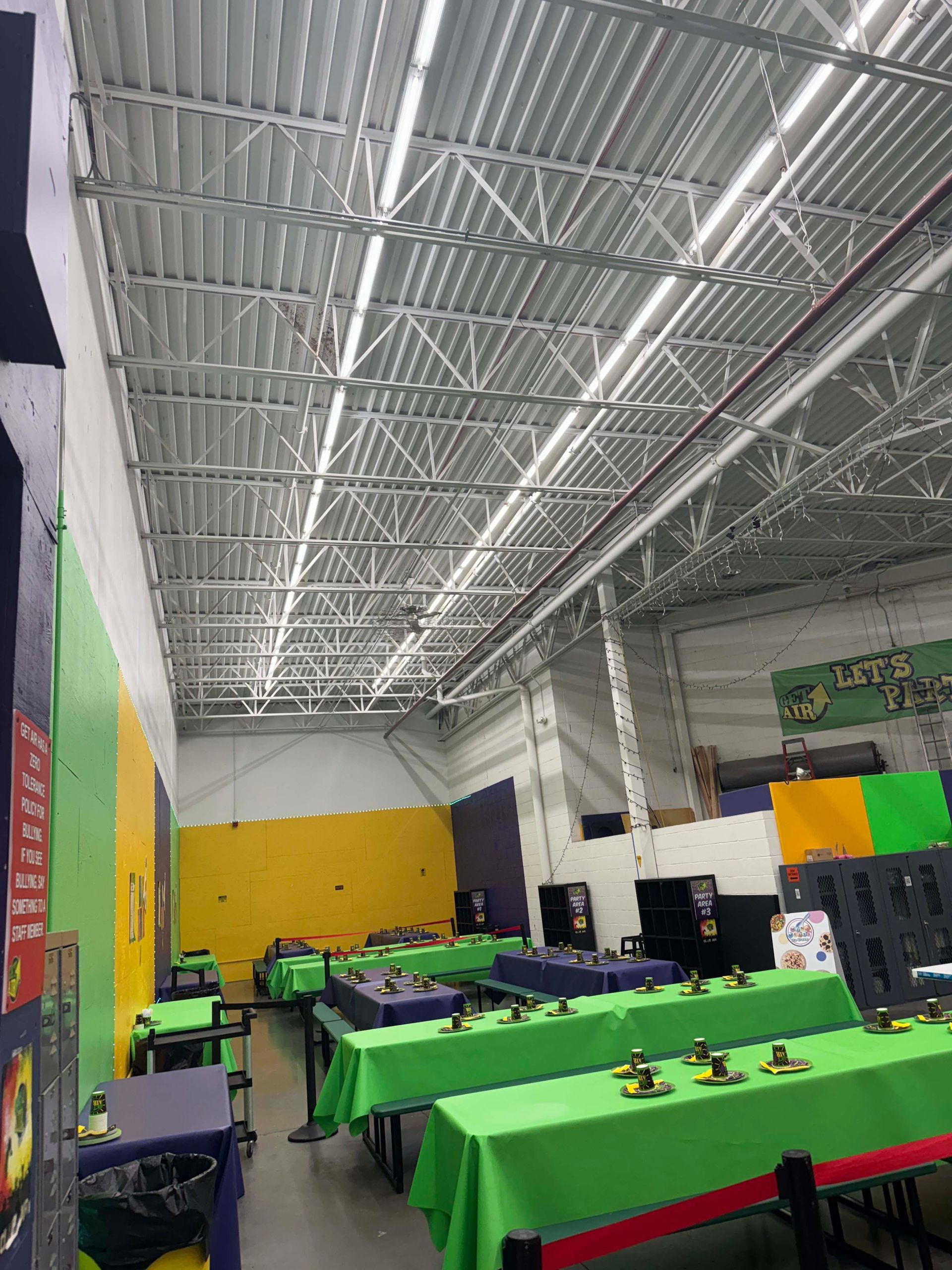 Indoor event hall with green-covered tables, metal truss ceiling, and colorful wall panels