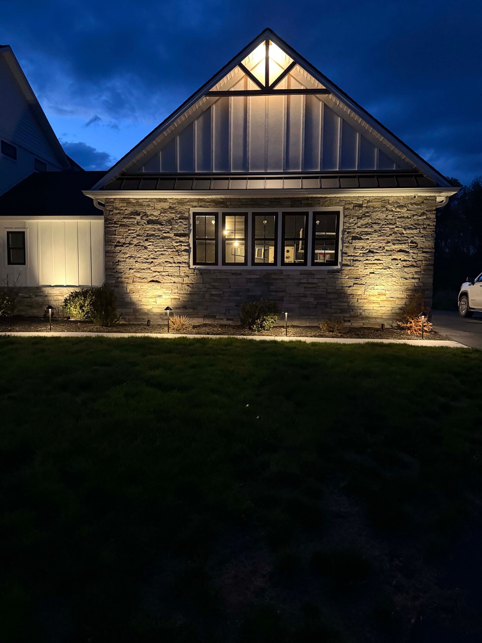 Stone house at dusk with lit front windows and warm exterior lighting under a blue evening sky