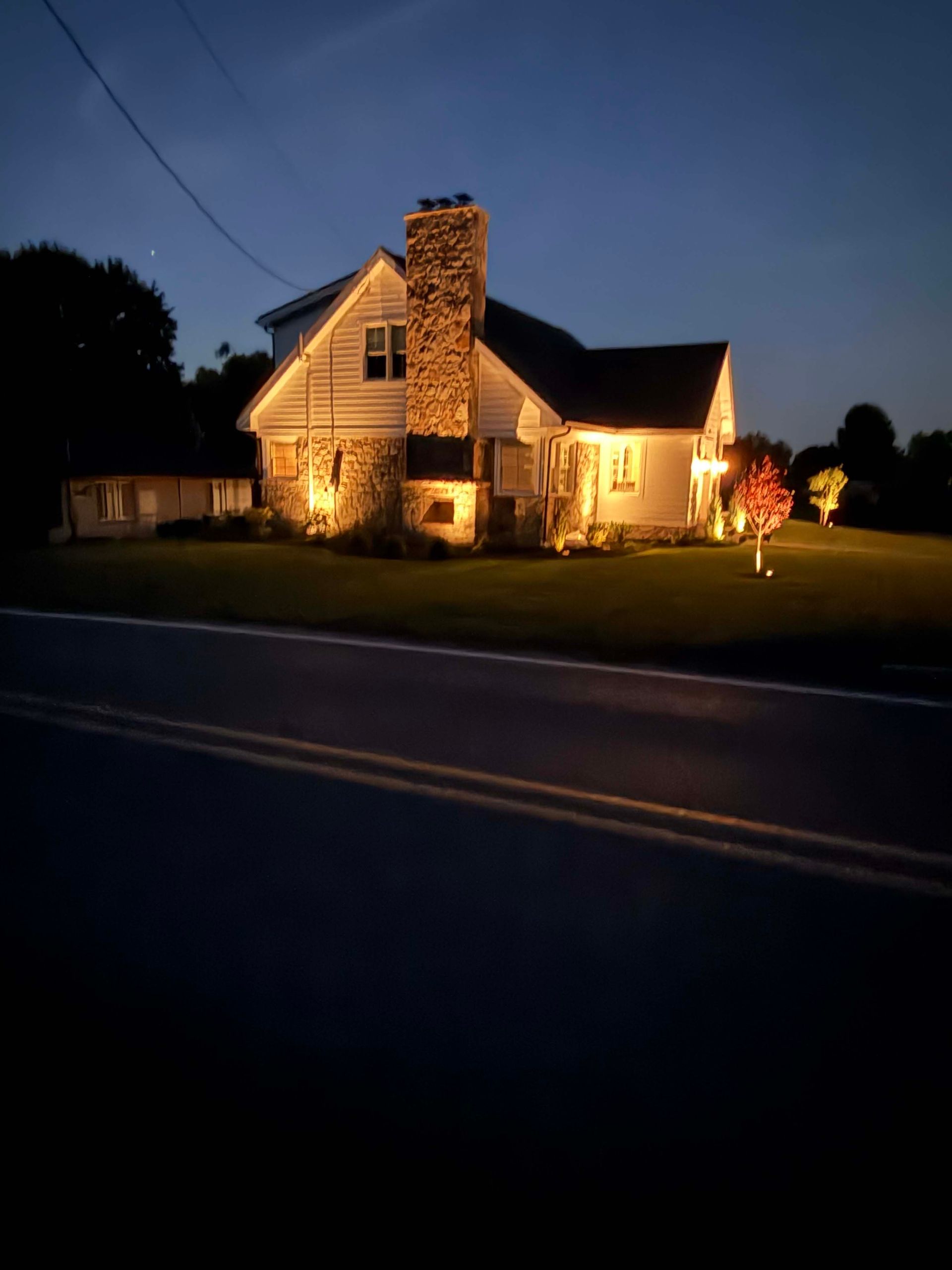 Lit house with stone chimney at dusk beside a road