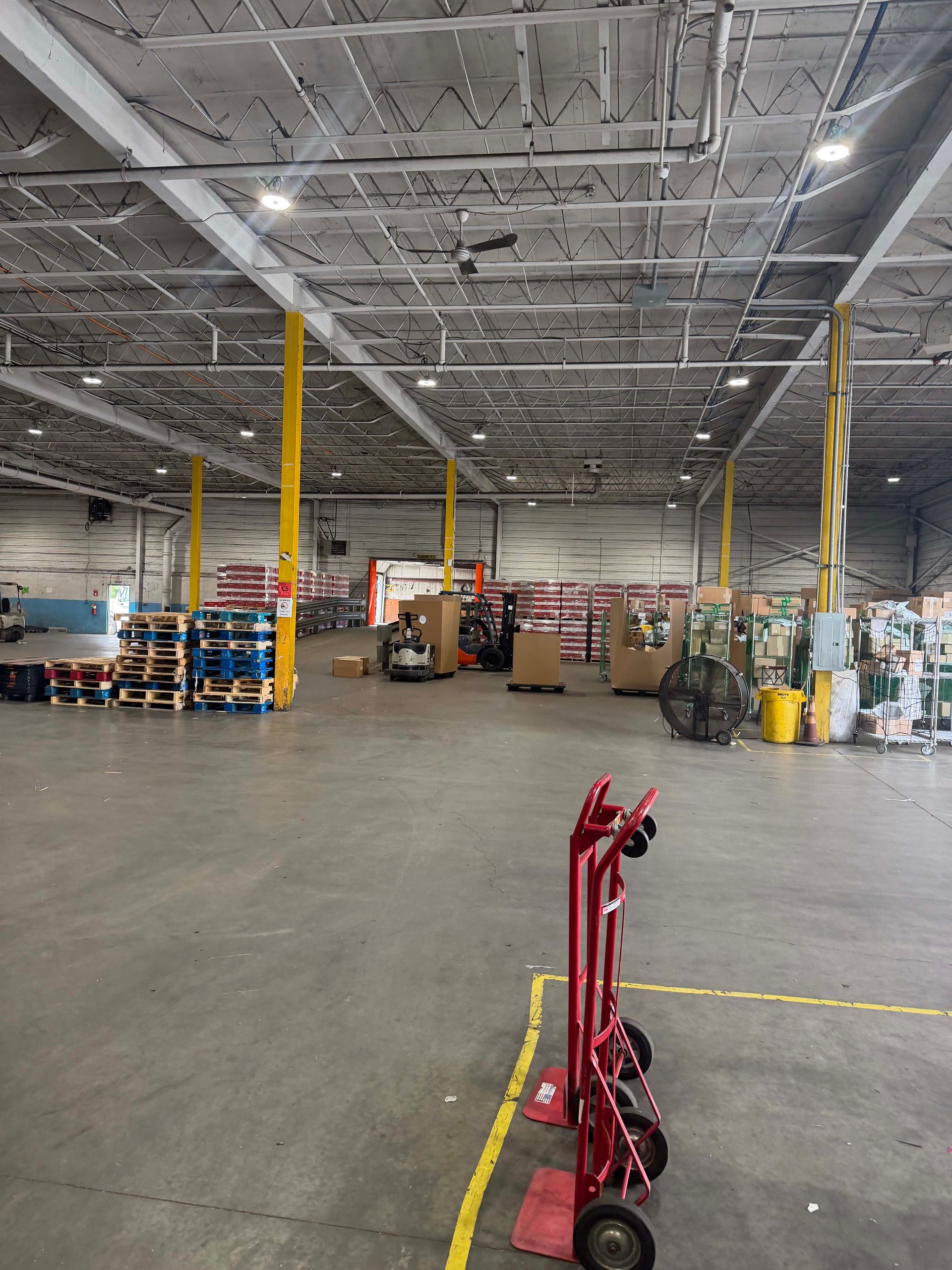 Warehouse interior with high ceiling, yellow columns, stacked pallets, and a red pallet jack in the foreground