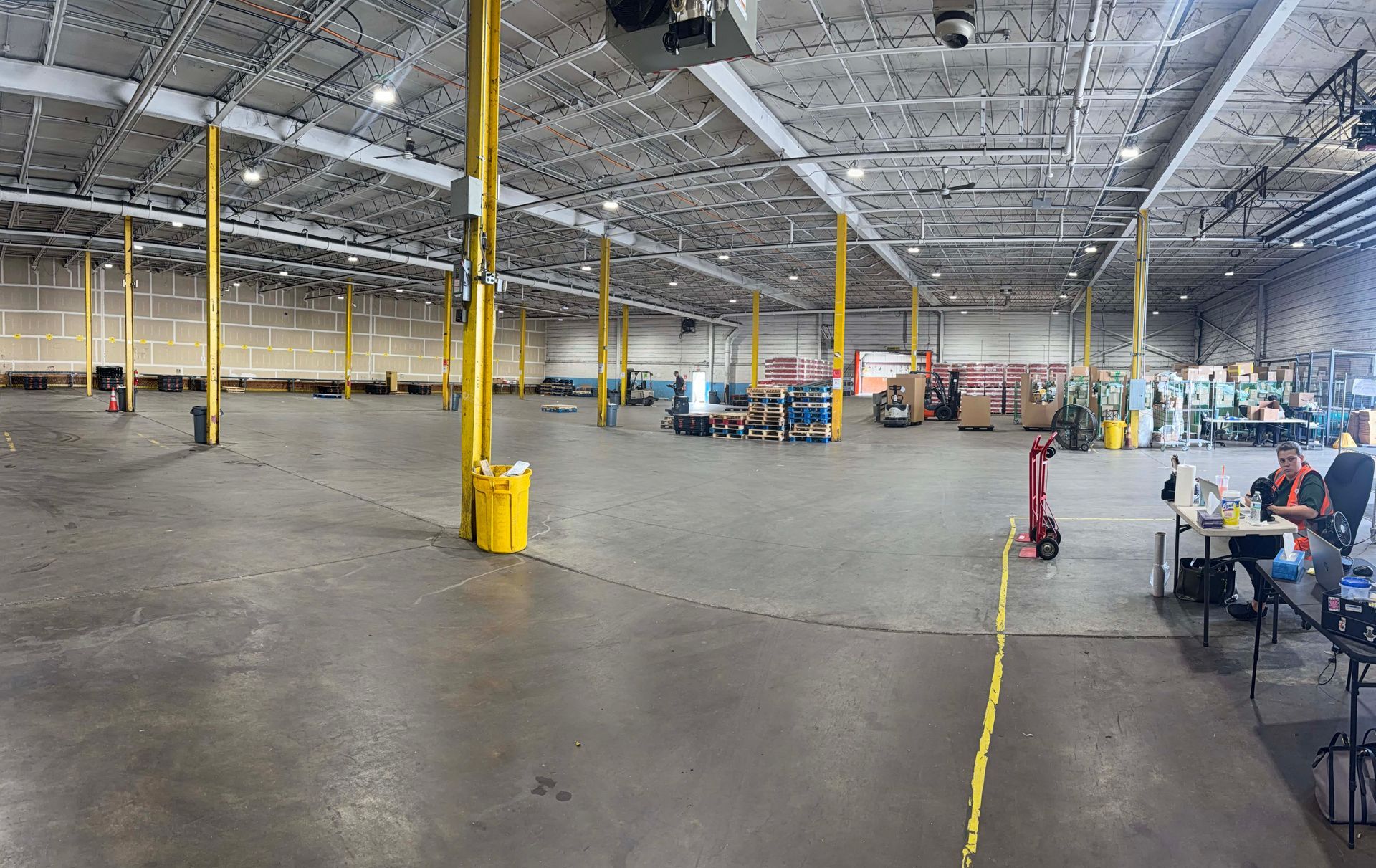Wide warehouse interior with high ceilings, yellow posts, and people seated at tables on the right
