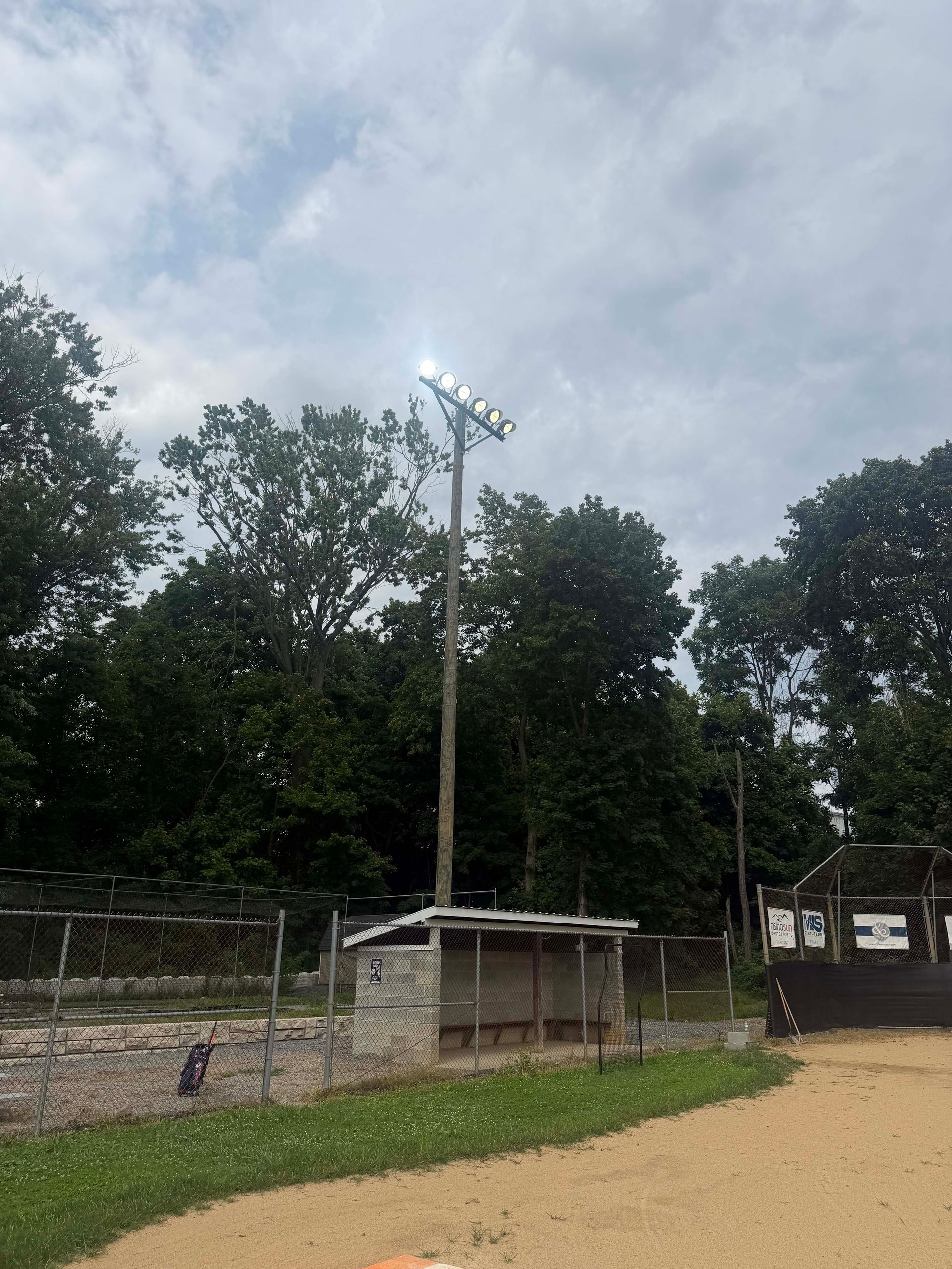 Lit sports field with tall floodlight pole, trees, and a small dugout under a cloudy sky.