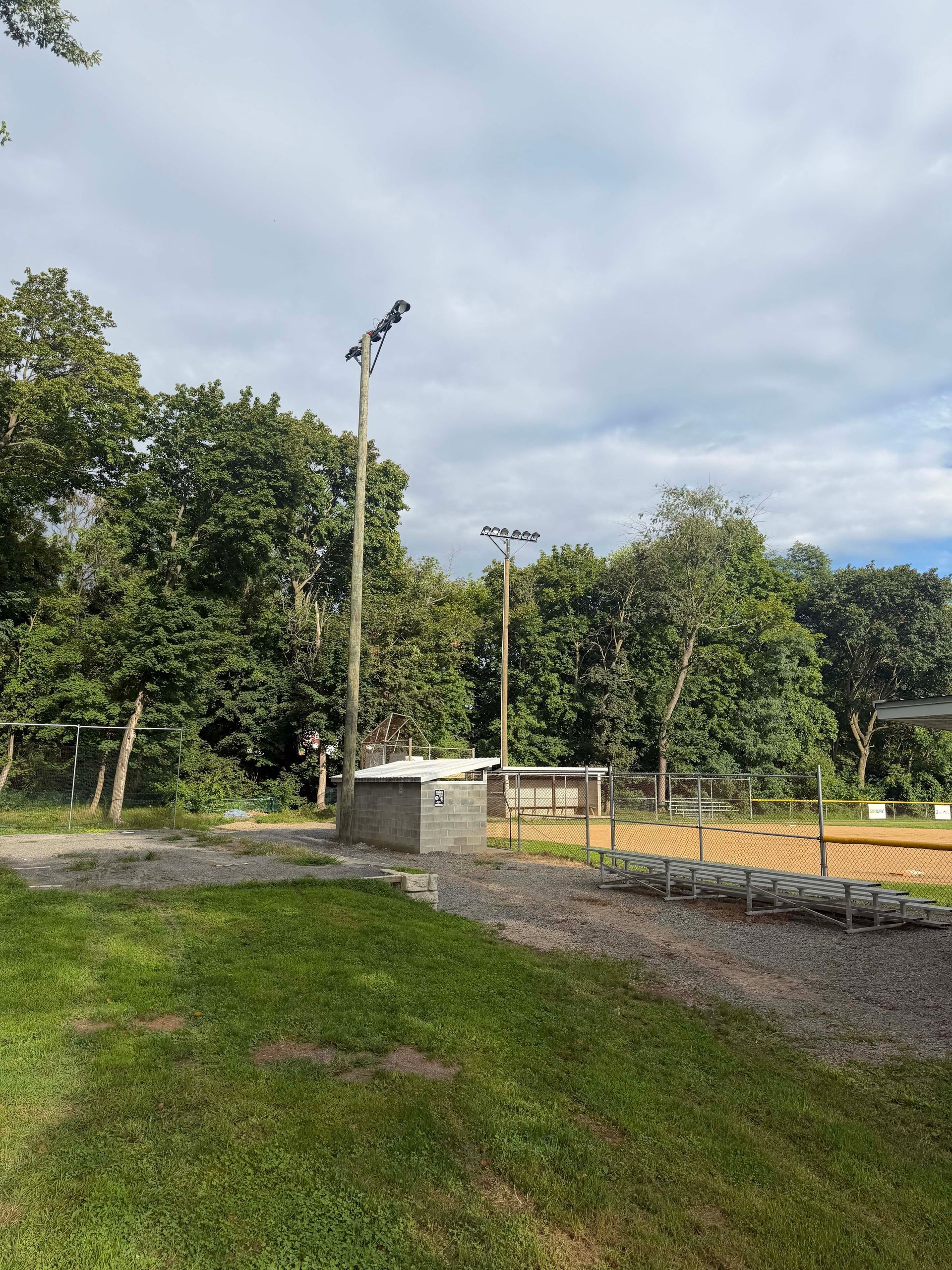 Grassy field with tall light poles, stone wall, trees, and cloudy sky