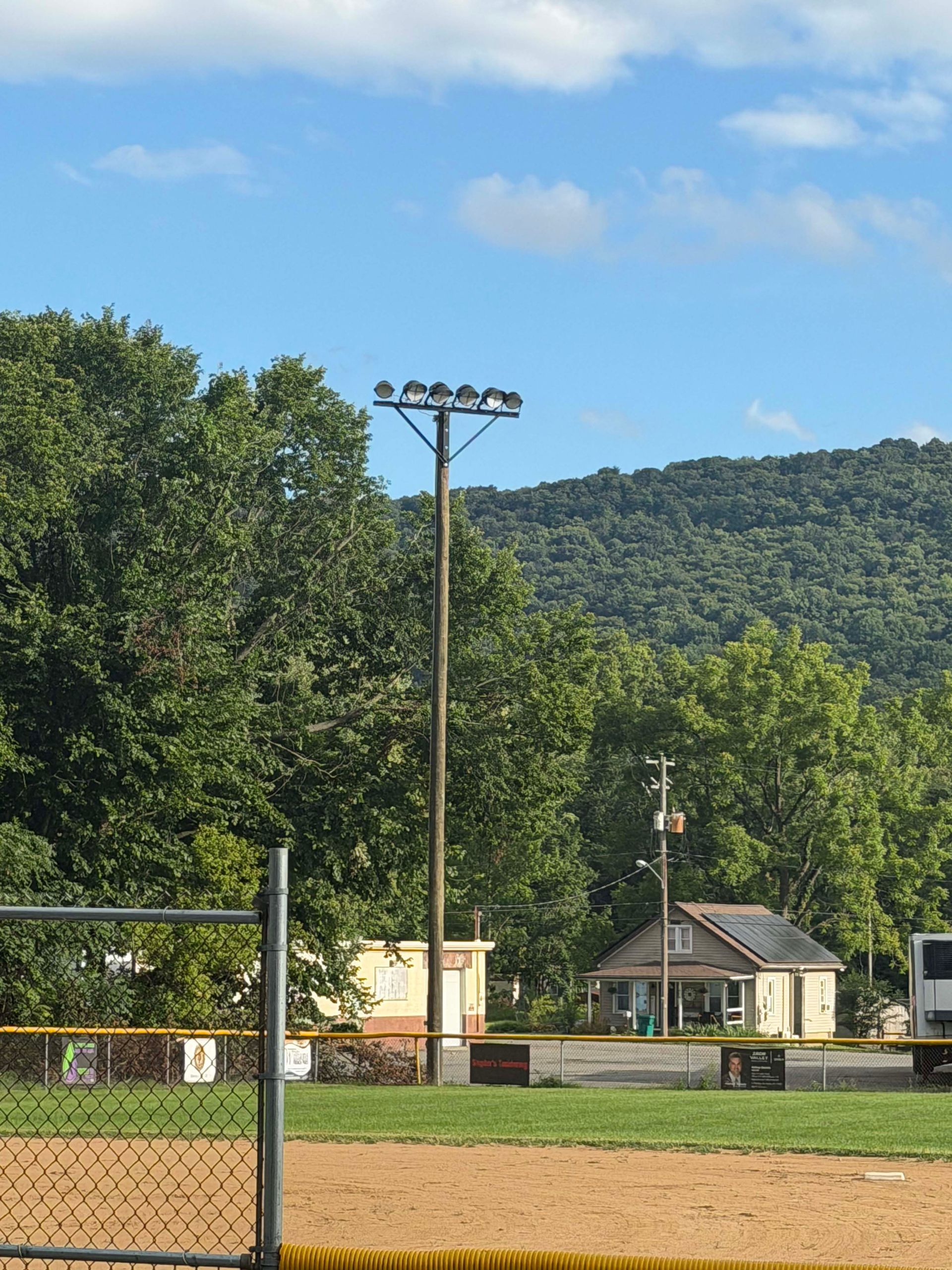Baseball field with a chain-link fence, light poles, and wooded hills under a blue sky