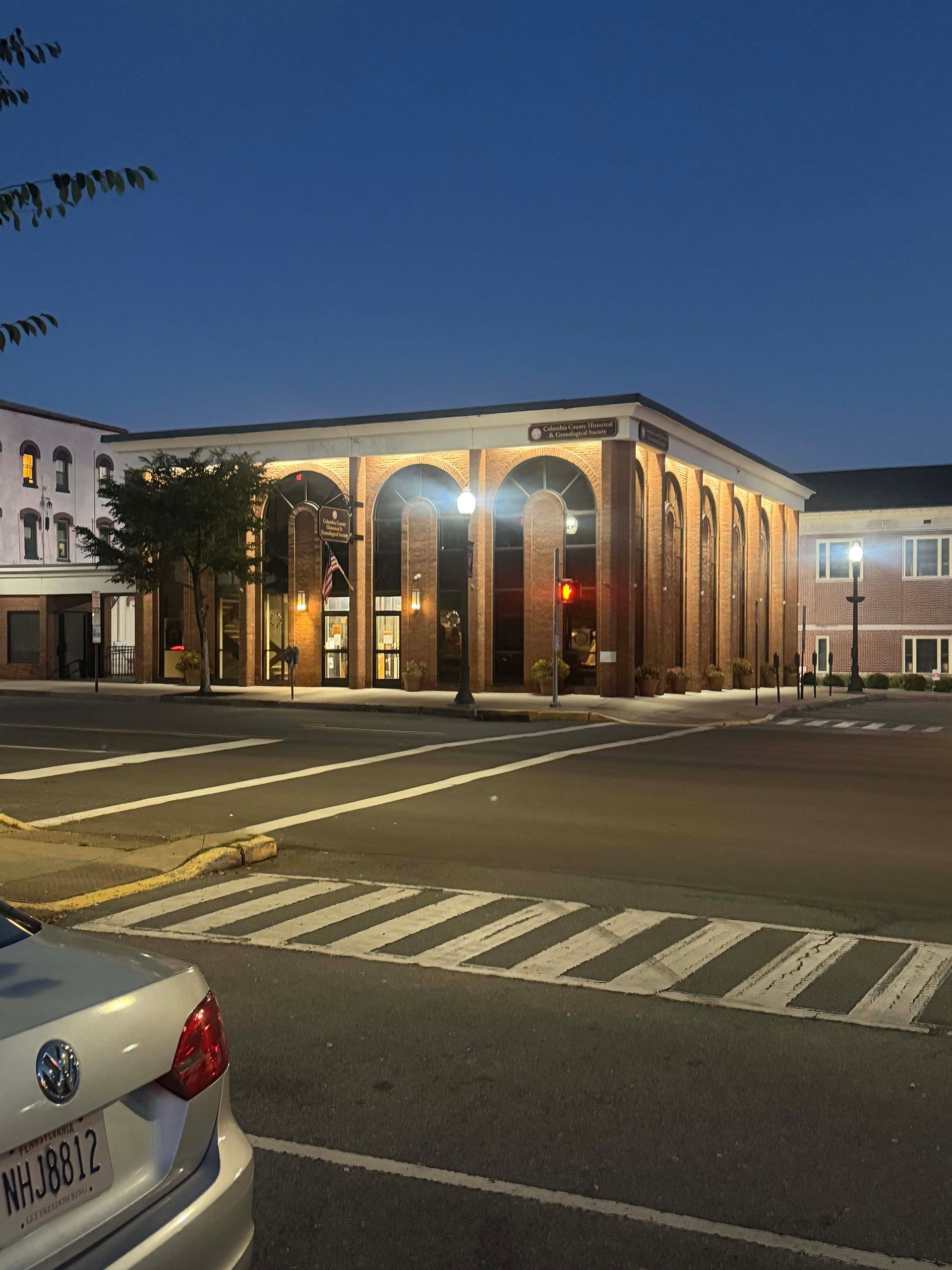 Lit brick building at dusk with lit arches, across a street and crosswalk.