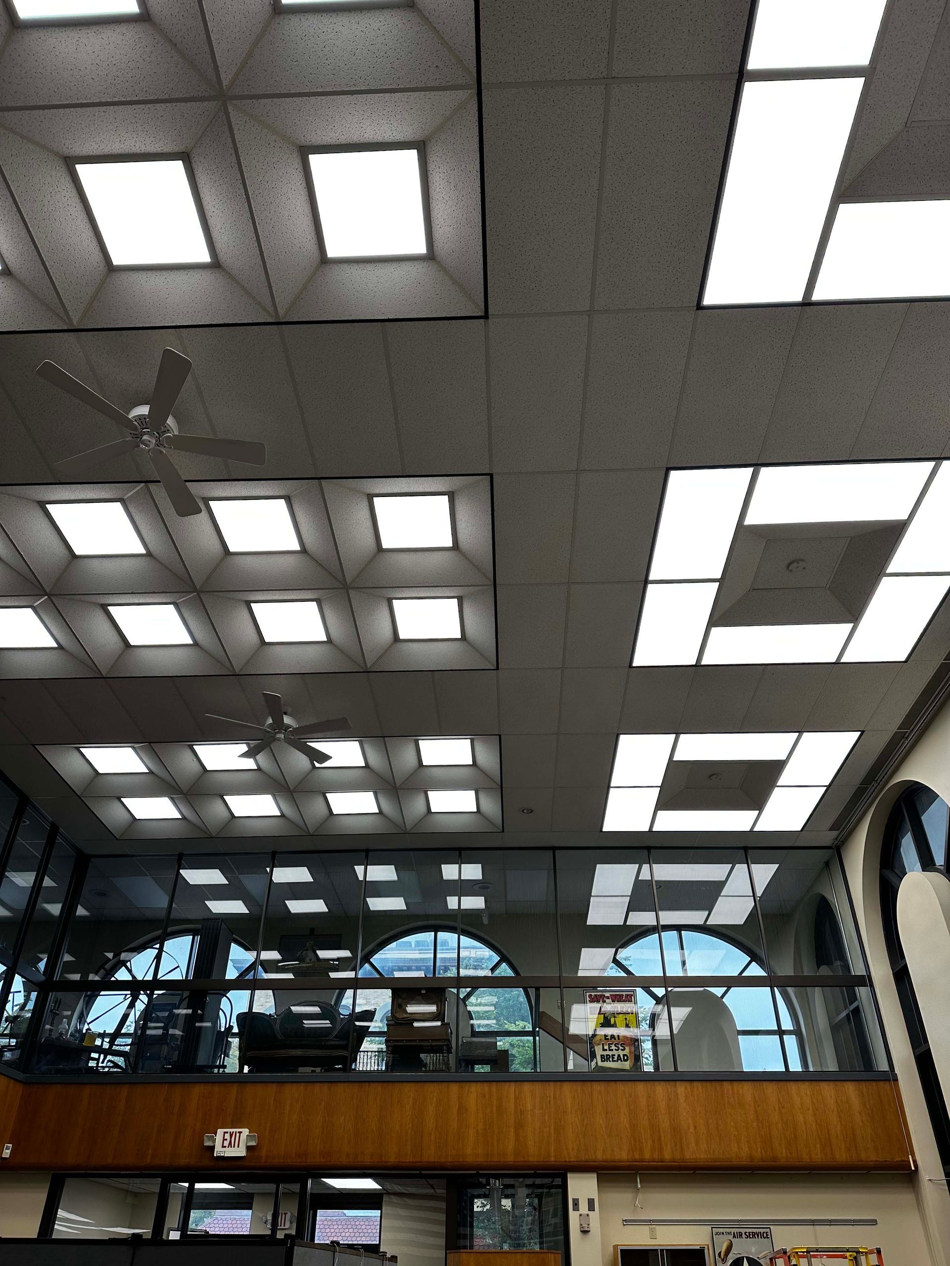Indoor lobby with geometric skylights, exposed beams, and a glass wall above a wooden balcony.