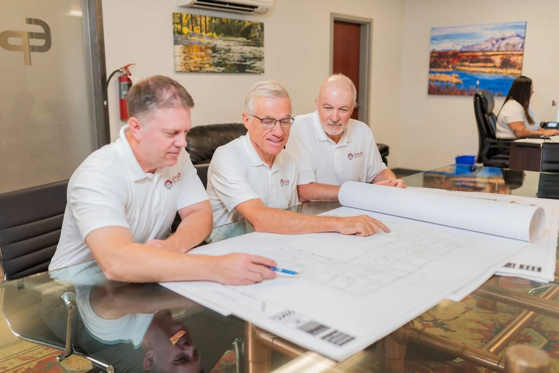 Three men in white polo shirts review blueprints at a conference table in an office.