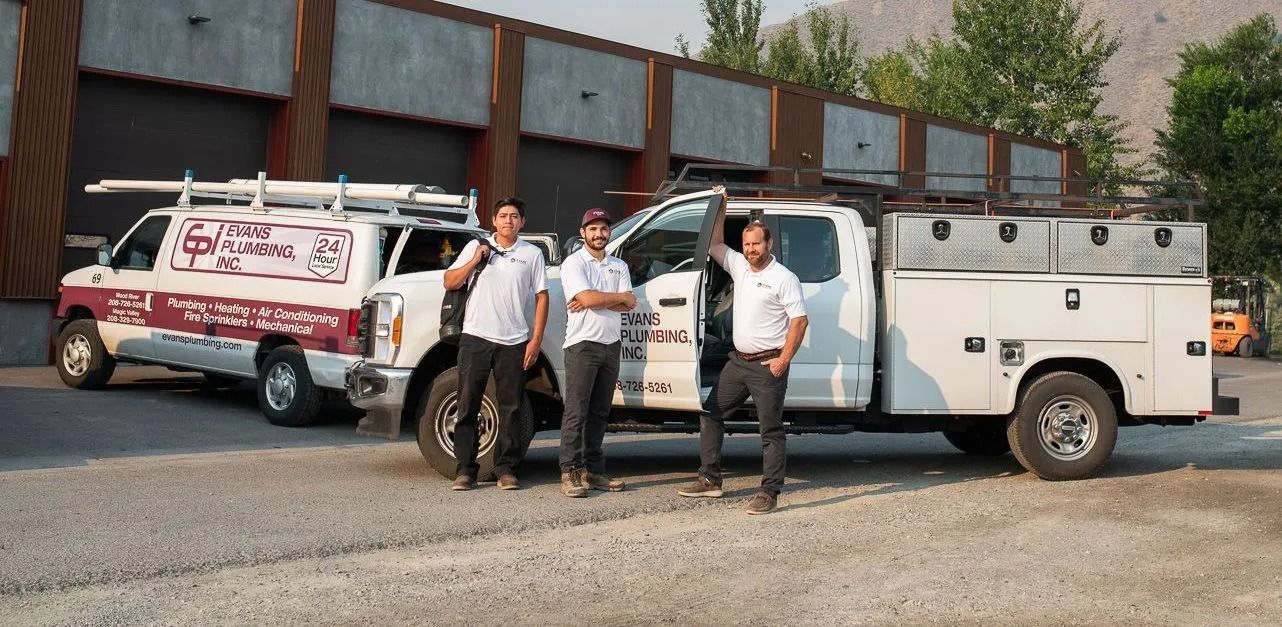 Three service workers stand smiling in front of two white work trucks parked in a gravel lot by a building.