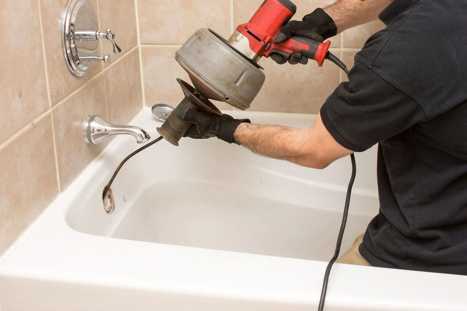 A plumber in a black shirt uses an electric drain snake to clear a clog in a white bathtub.