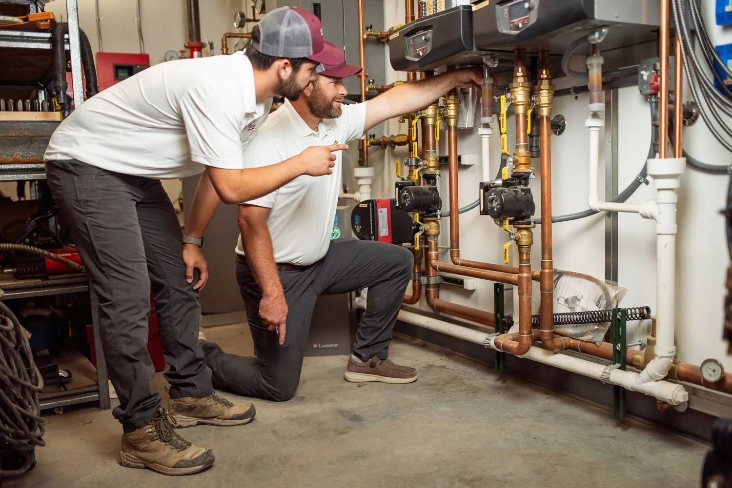 Two technicians in matching uniforms inspect and discuss complex industrial plumbing and heating equipment.