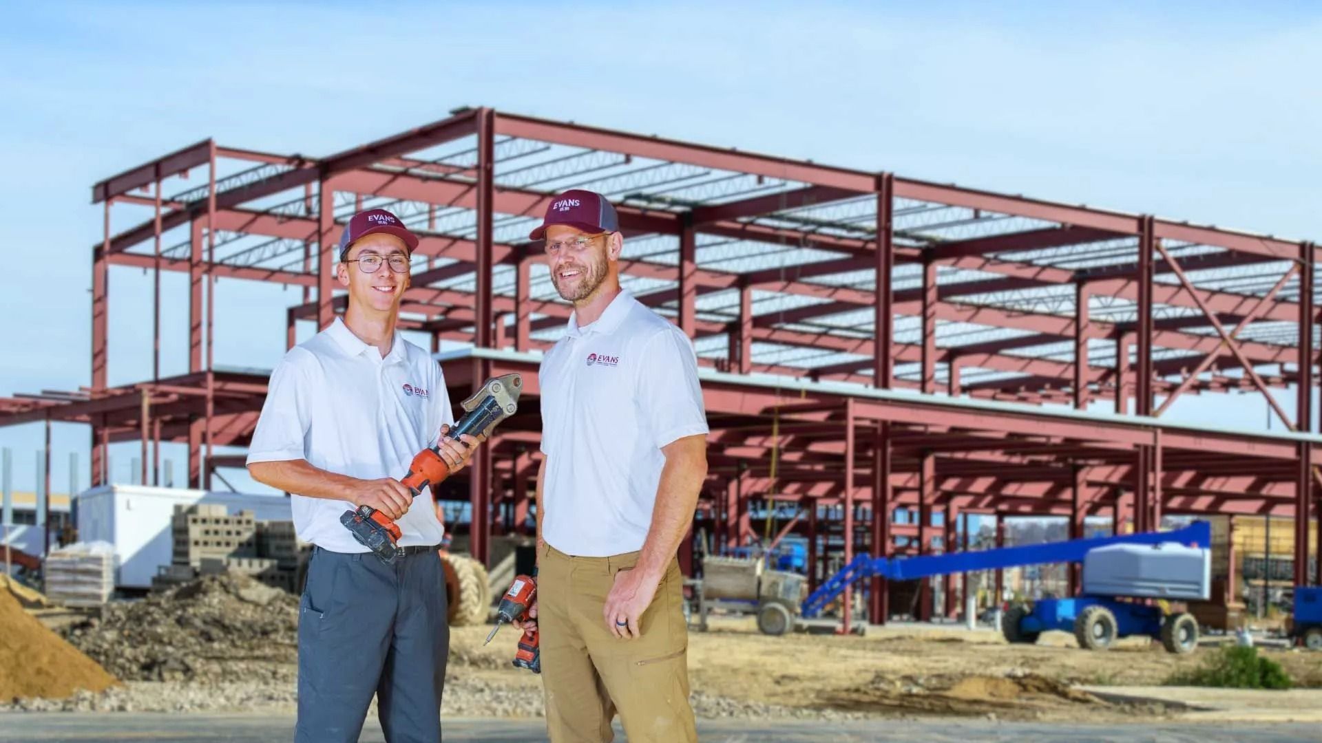 Two people in matching hats and white shirts stand before a large red steel construction framework on a sunny day.