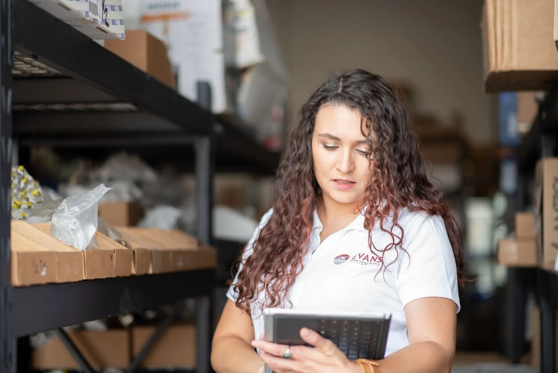 A person with long curly hair wearing a white polo shirt reviews data on a tablet in a warehouse filled with boxes.