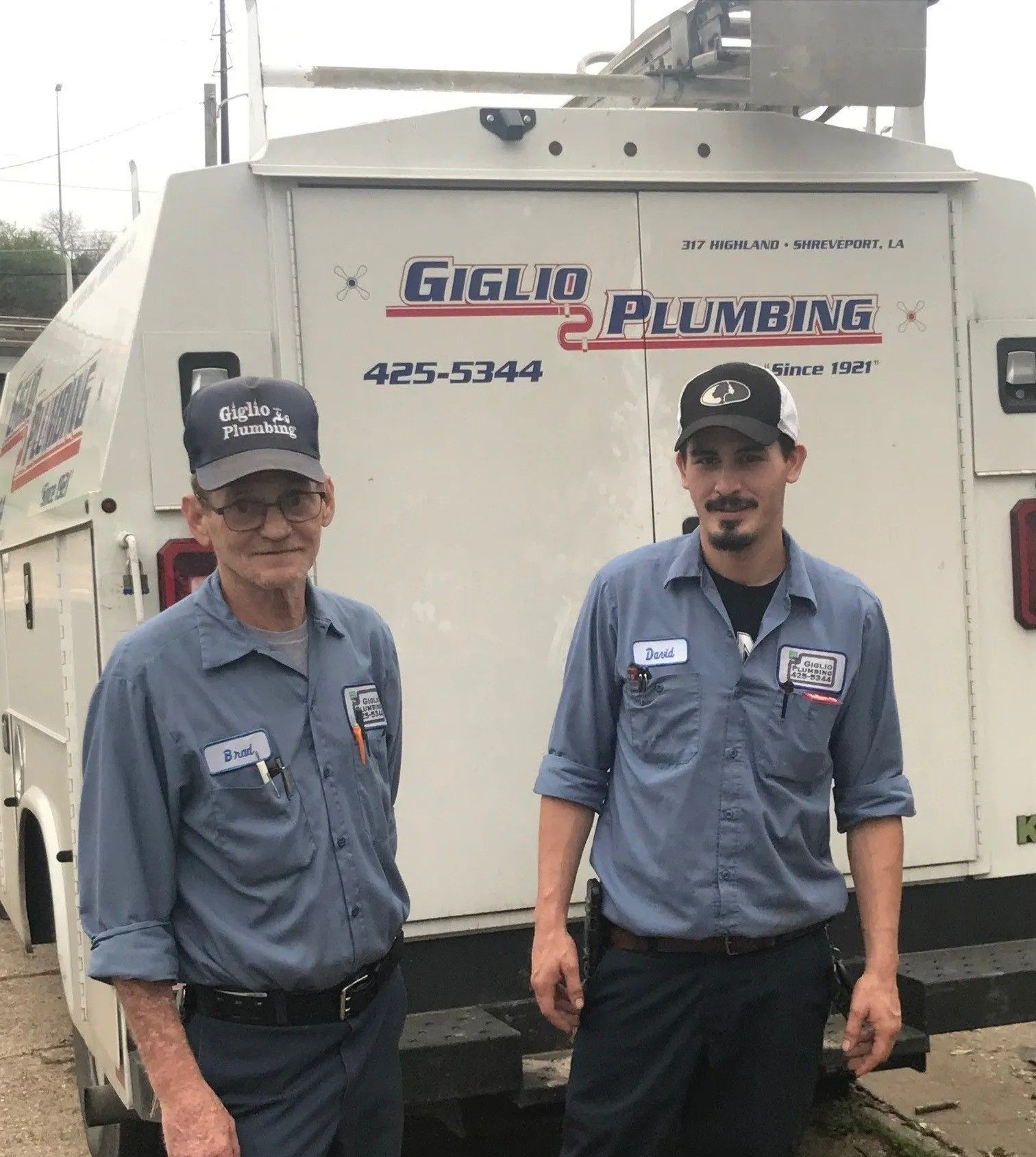 Two plumbers standing in front of a white service truck with “Plumbing” signage.