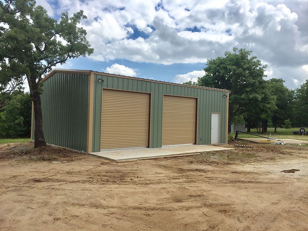 A green metal building with two garage doors is in a dirt field.