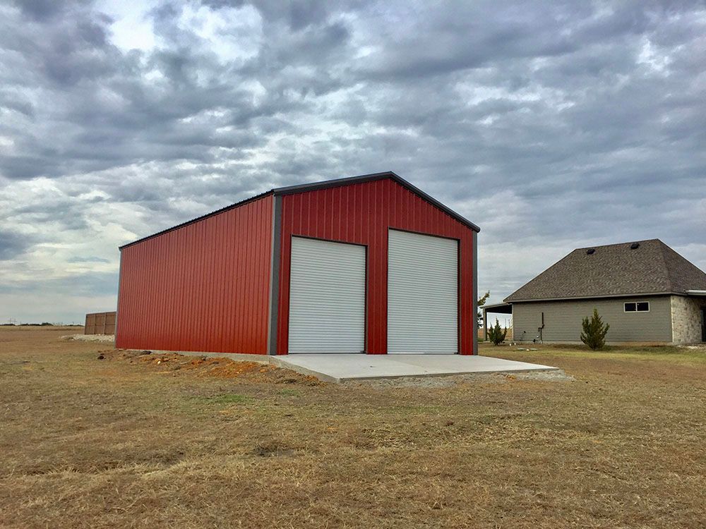 A red metal building with two white doors is in a field next to a house.