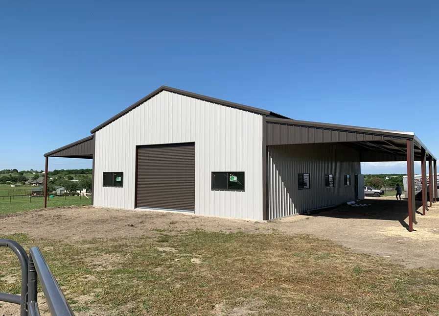 A large white metal building with a brown roof is sitting in the middle of a grassy field.