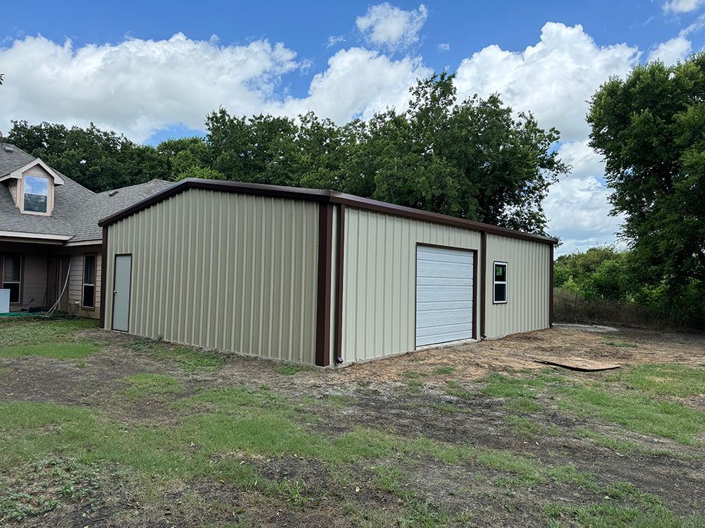 A large metal garage is sitting in the middle of a grassy field next to a house.