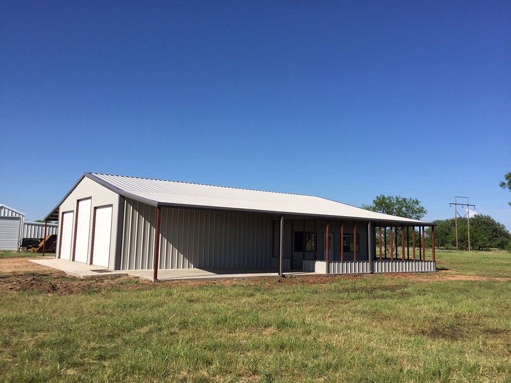 A large metal building with a porch is sitting in the middle of a grassy field.