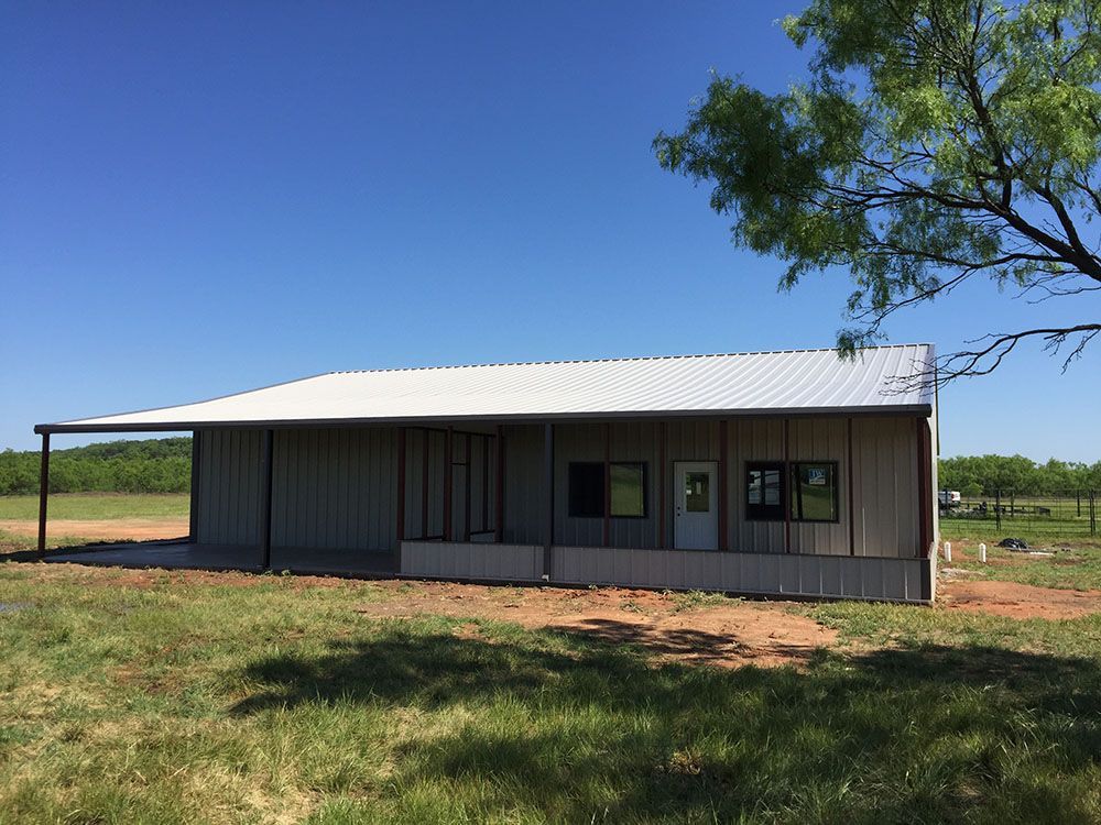 A large metal building with a porch is sitting in the middle of a grassy field.