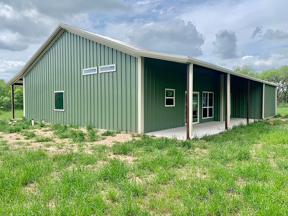 A green metal building with a porch is sitting in the middle of a grassy field.