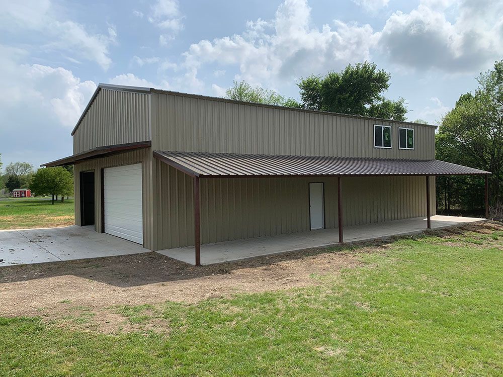A large metal building with a porch and a garage door is sitting in the middle of a grassy field.