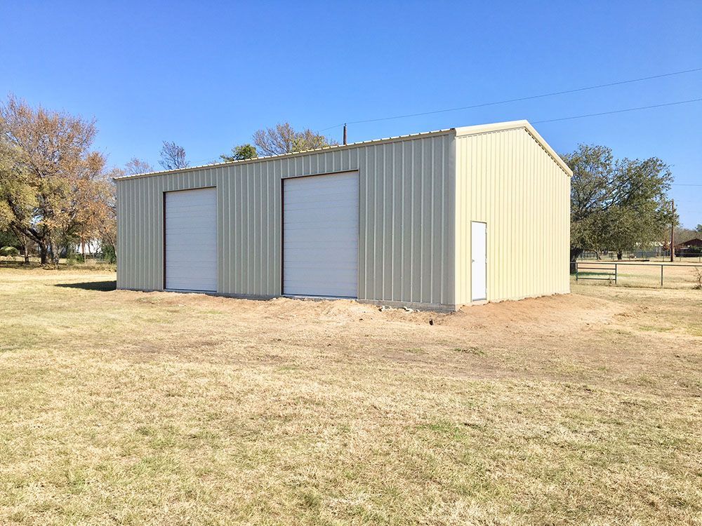 A metal building with two garage doors is sitting in the middle of a grassy field.