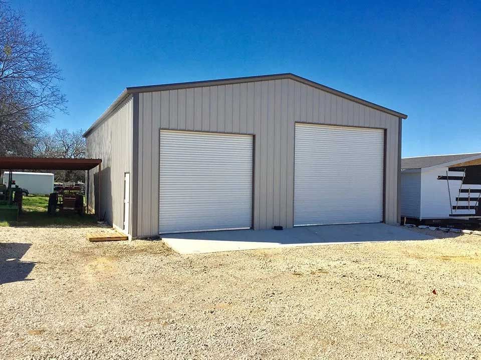 A garage with two white garage doors is sitting on top of a gravel lot.