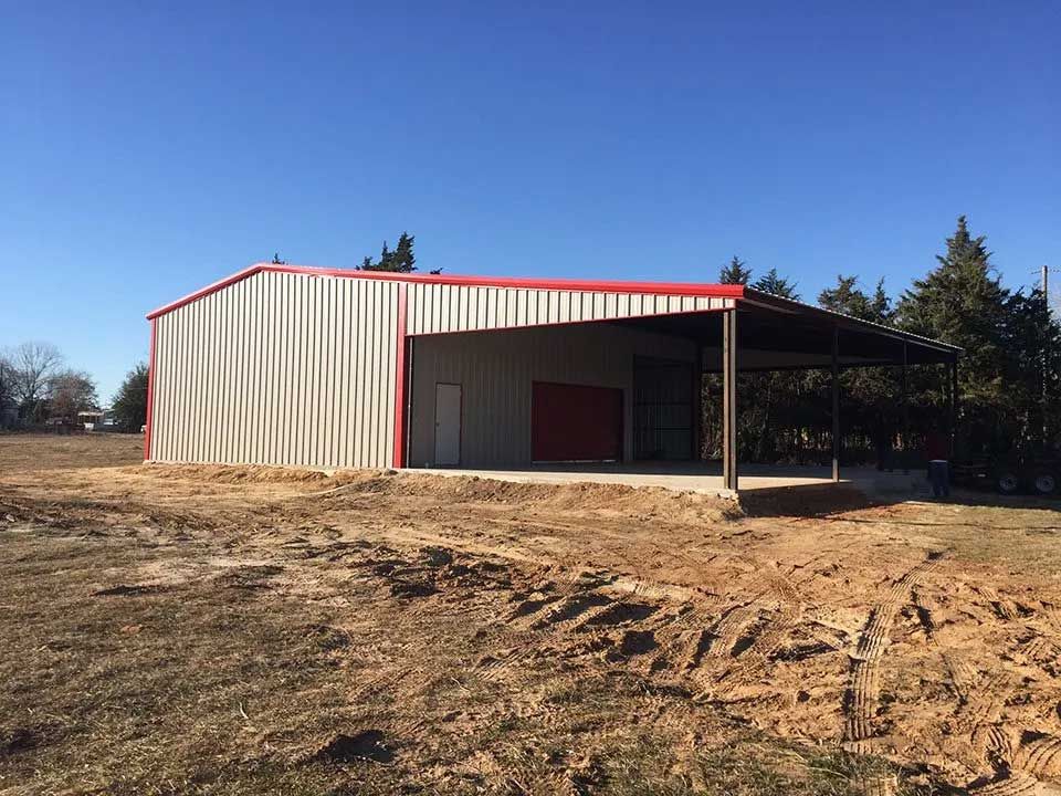 A large metal building with a red roof is sitting in the middle of a dirt field.