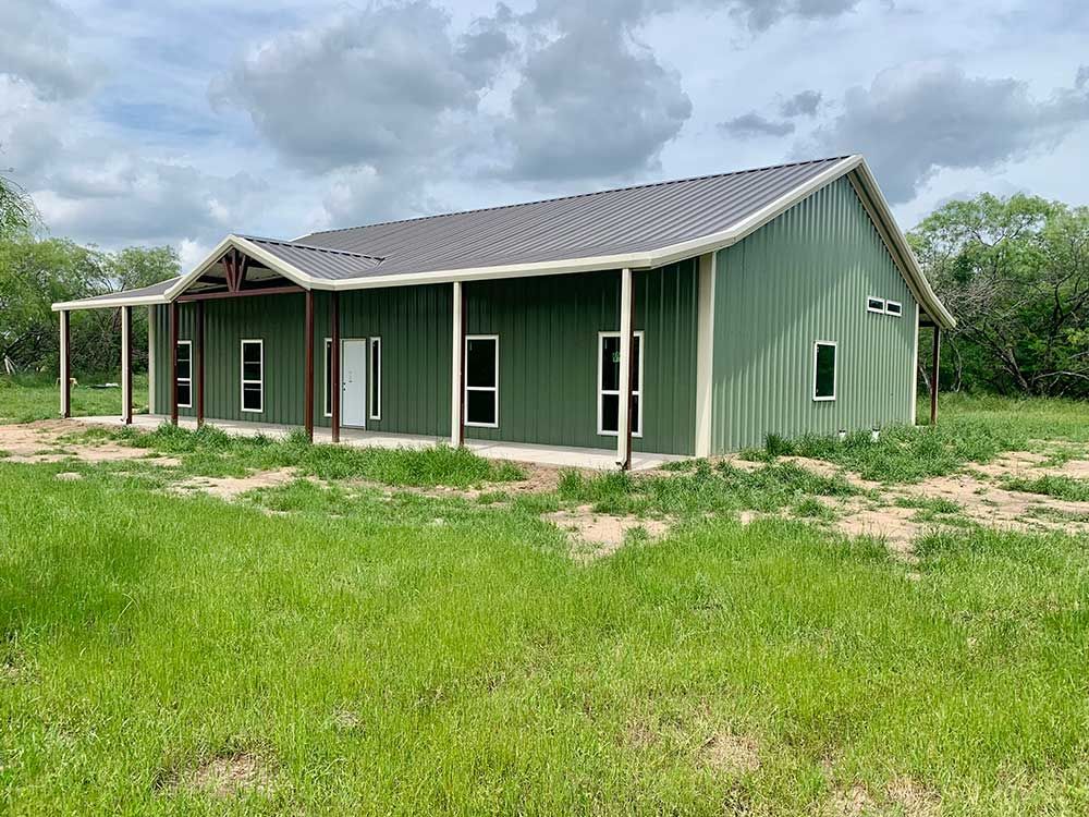 A green metal building with a porch is sitting in the middle of a grassy field.