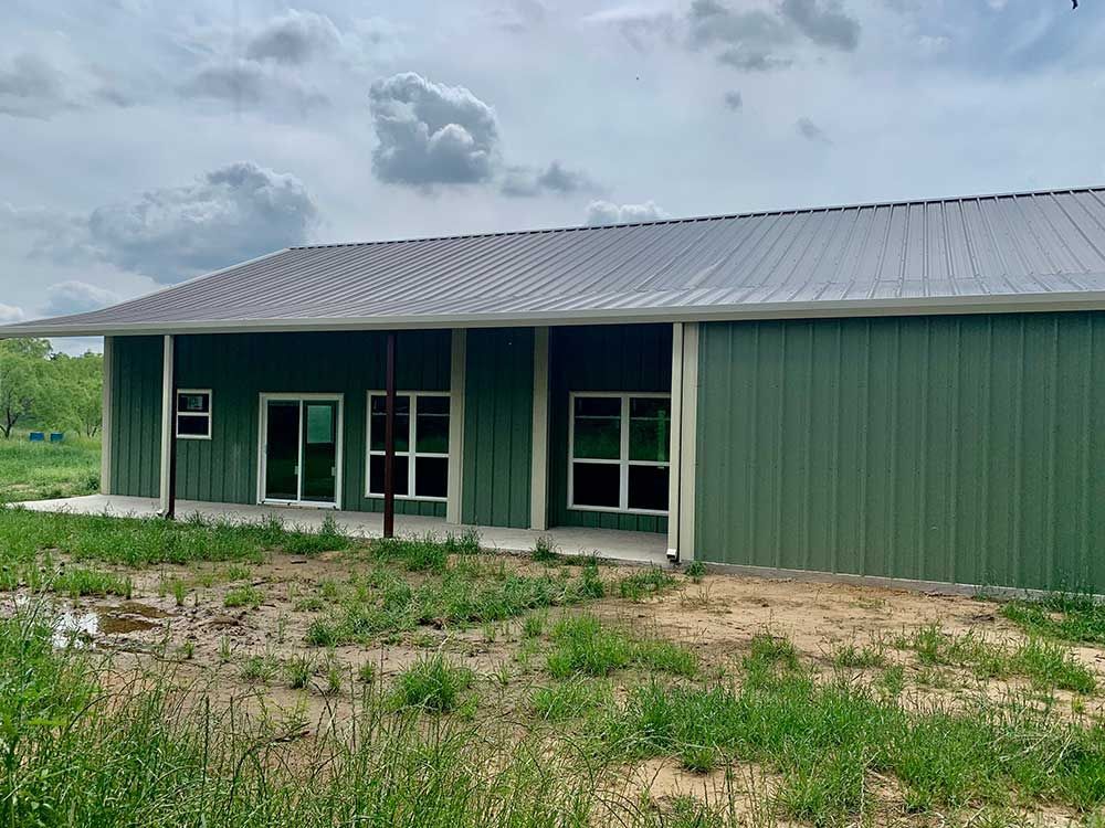 A large green building with a metal roof is sitting in the middle of a grassy field.