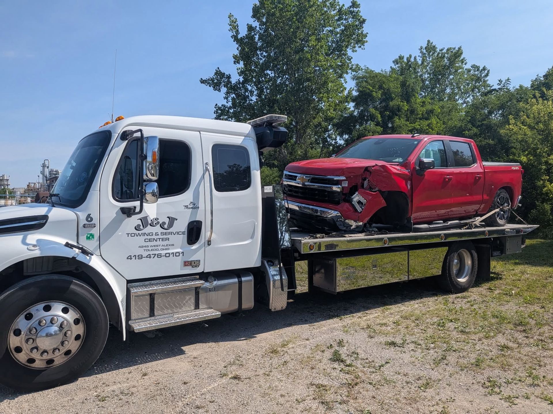 Tow truck carrying a damaged red pickup truck on a flatbed, parked outdoors under a blue sky.